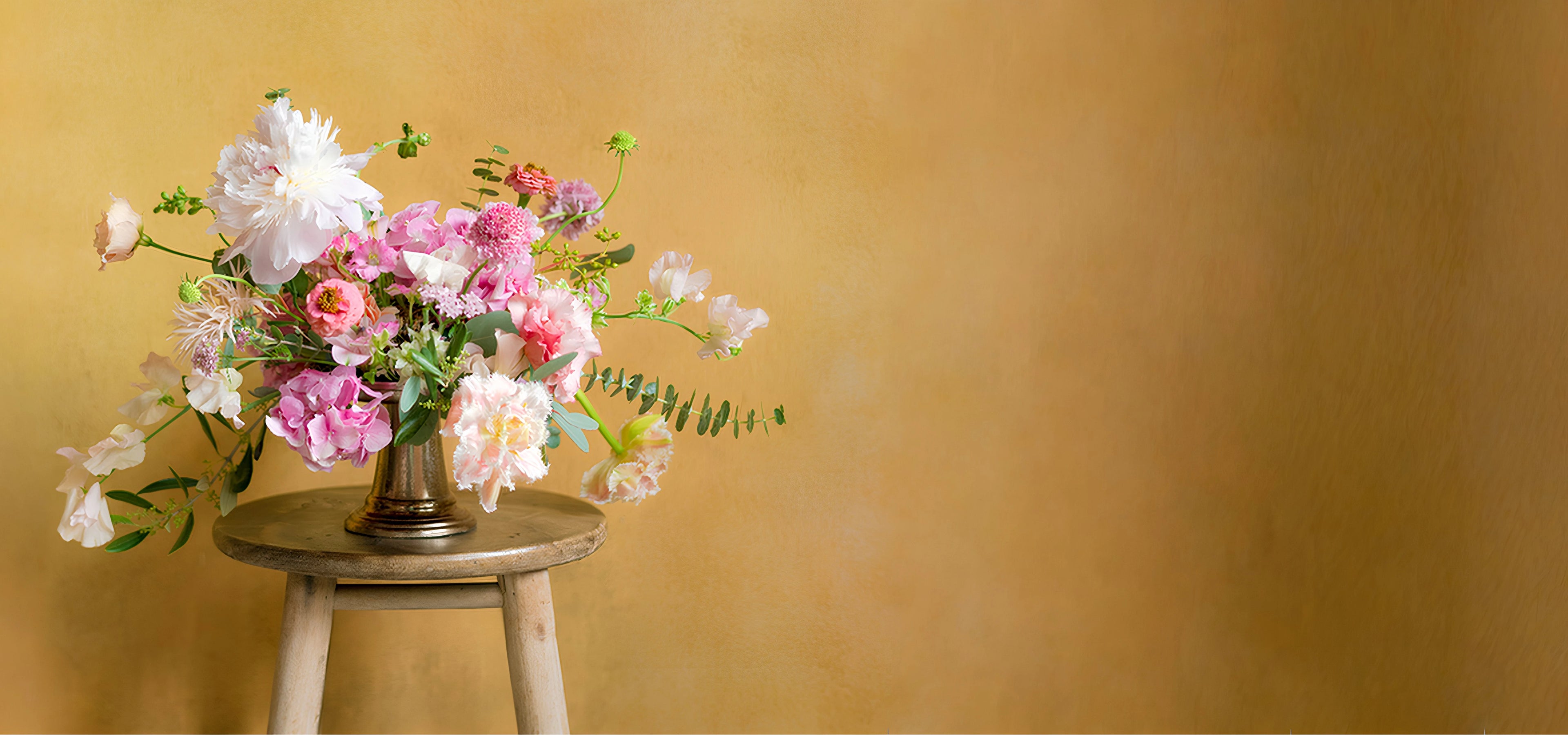Bouquets of flowers in a vase on top of a stool against a yellow background.