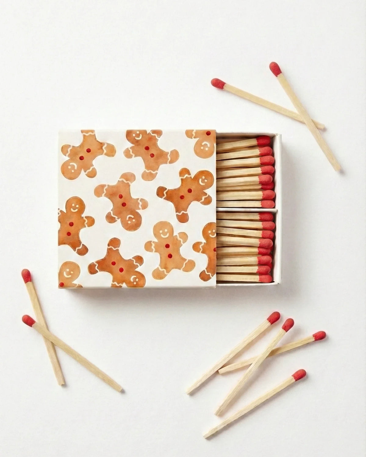 A matchbox with a white lid decorated with gingerbread men illustrations, shown open with wooden matches and red tips scattered around on a white background.