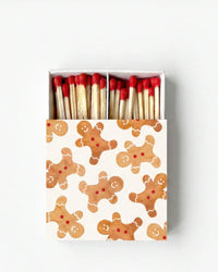 A matchbox filled with wooden matches topped with red tips, shown from above on a light gray background. The box lid has a white base printed with a repeating pattern of gingerbread cookies in warm brown tones.