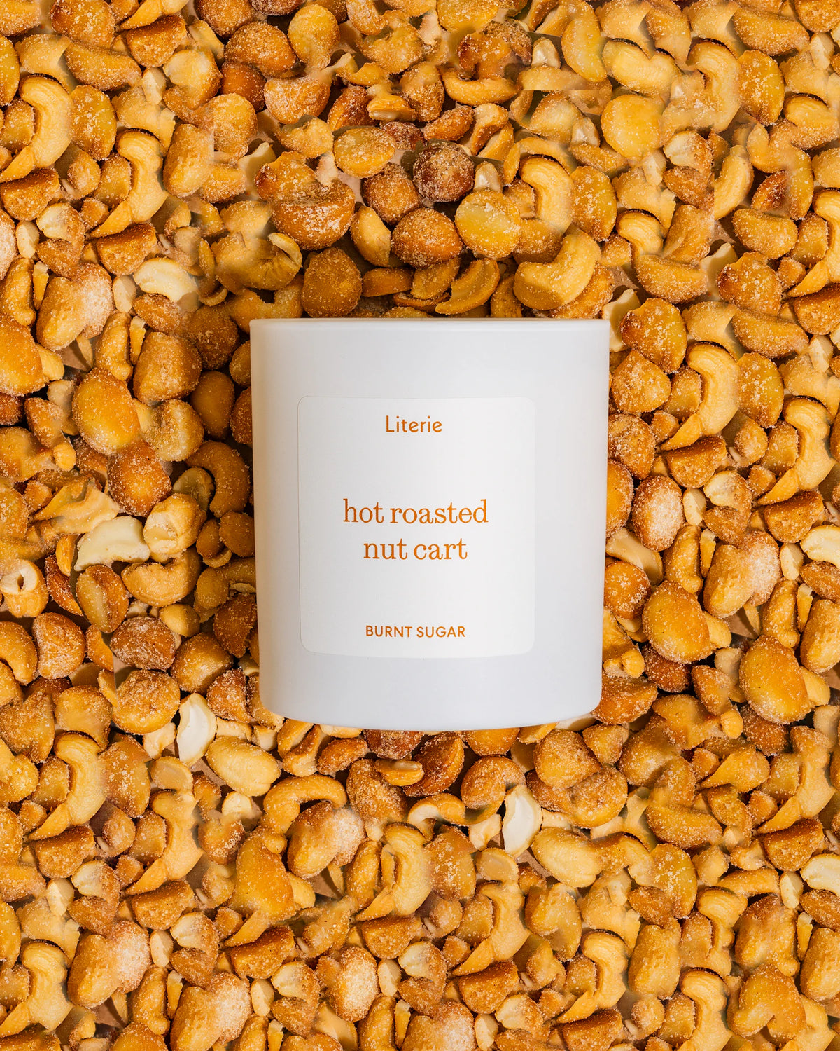 A white candle jar labeled 'Literie hot roasted nut cart' and 'BURNT SUGAR' sits centered on a dense background of golden-brown roasted cashews.