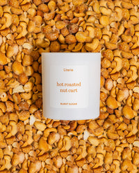A white candle jar labeled 'Literie hot roasted nut cart' and 'BURNT SUGAR' sits centered on a dense background of golden-brown roasted cashews.