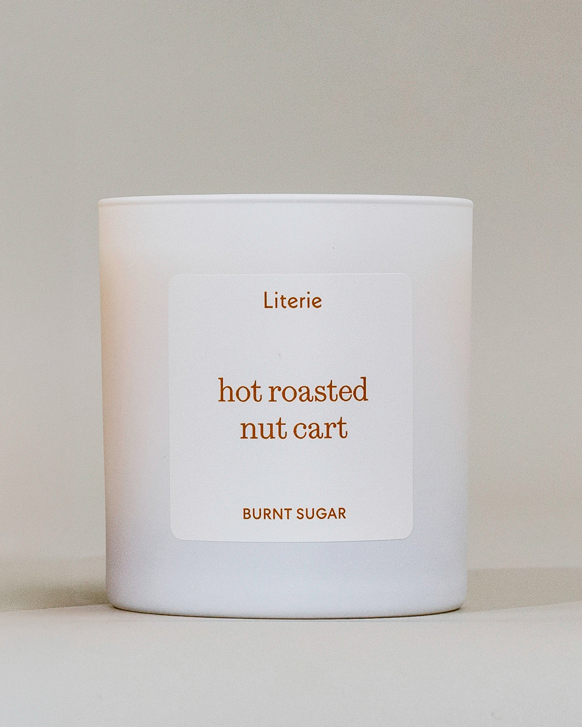 A white candle jar with a minimalist white label featuring the scent name 'hot roasted nut cart' and brand 'Literie' in a warm brown color, set against a neutral beige background.