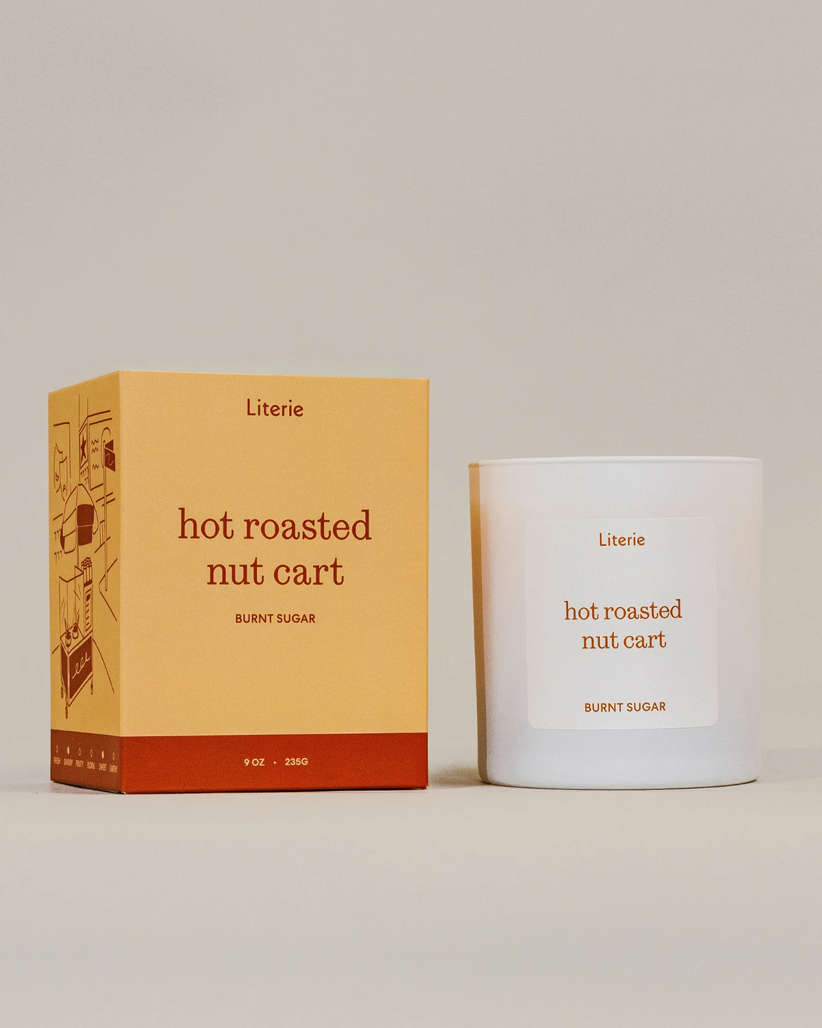 A white glass candle jar with a white label next to its mustard yellow and rust-colored packaging box. Both items feature the brand name 'Literie' and the scent name 'hot roasted nut cart' on a clean, solid, warm beige background.