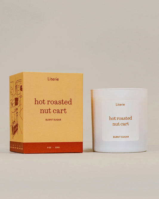 A white glass candle jar with a white label next to its mustard yellow and rust-colored packaging box. Both items feature the brand name 'Literie' and the scent name 'hot roasted nut cart' on a clean, solid, warm beige background.