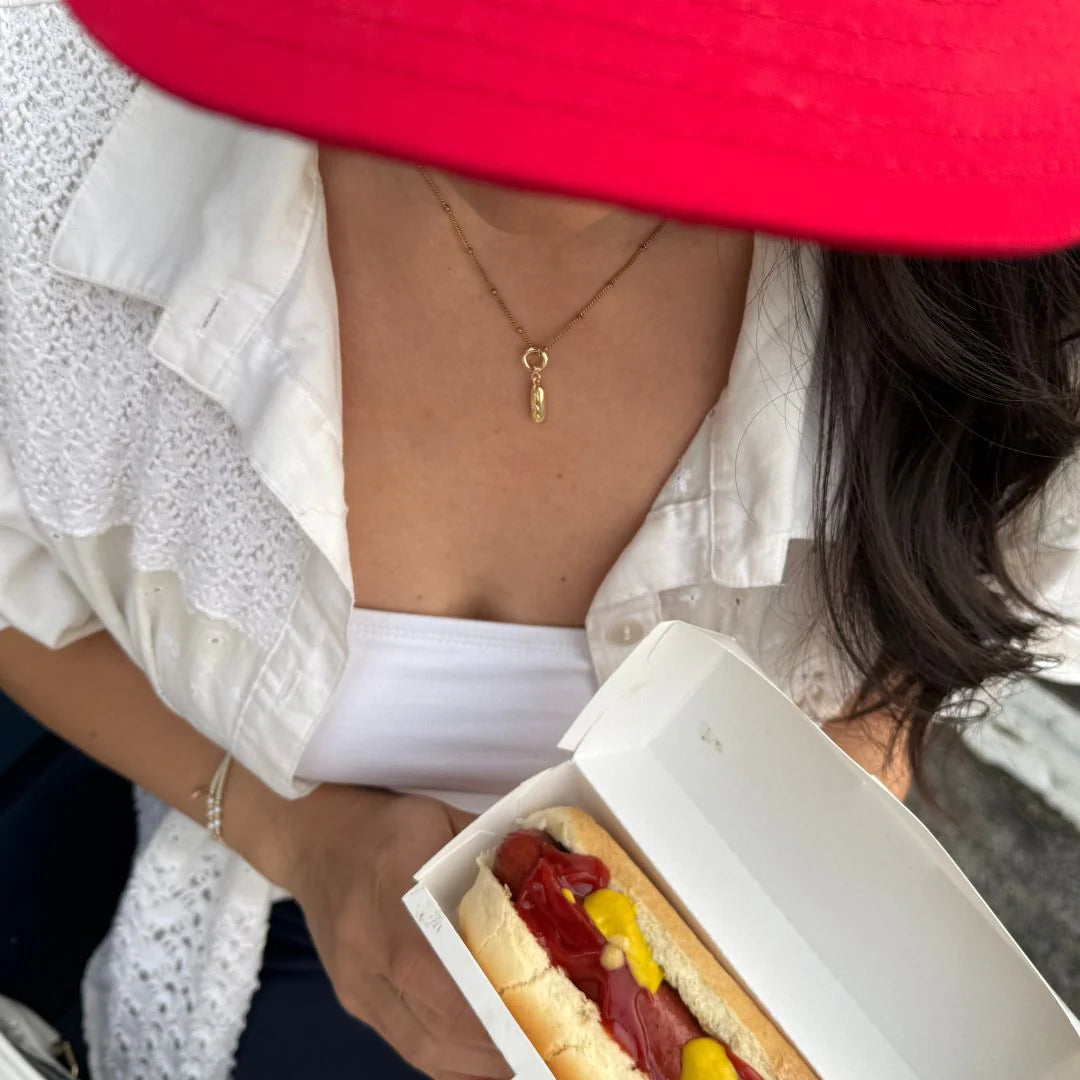 Hotdog charm on necklace with hotdog being held by a woman with a red hat.