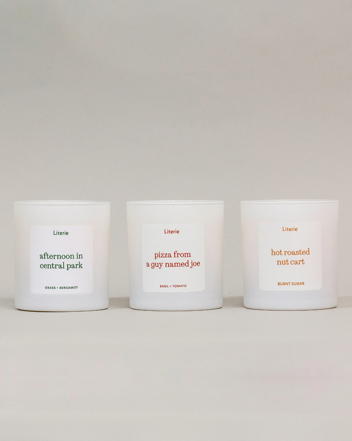 Three white scented candles in matte white jars, arranged side by side on a light gray surface against a neutral gray background. Each candle has a minimalist white label with black and muted red text, featuring the brand 'Literie' and scent names: 'afternoon in central park', 'pizza from a gay named joe', and 'hot roasted nut cart'.