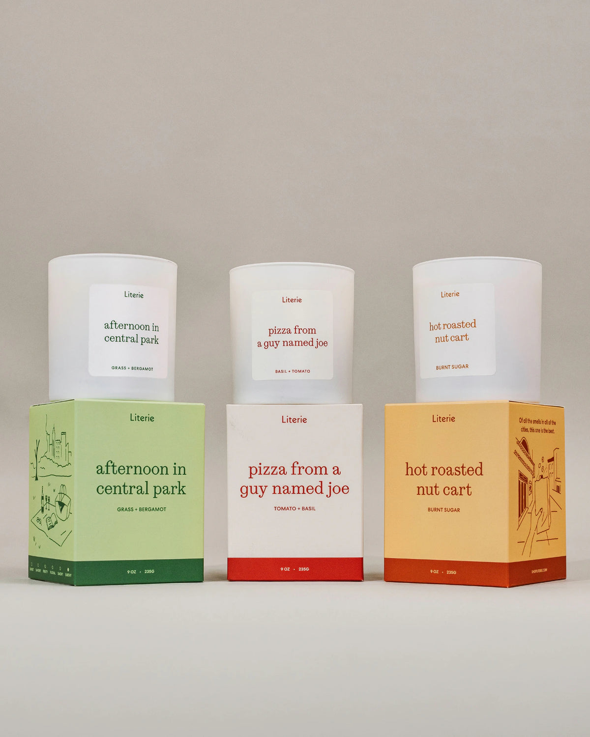 Three Literie scented candles in white glass with white labels, displayed alongside their colorful cardboard boxes on a neutral studio background. The boxes are colored light green, white with red, and light orange, matching the scents: 'afternoon in central park', 'pizza from a guy named joe', and 'hot roasted nut cart'.