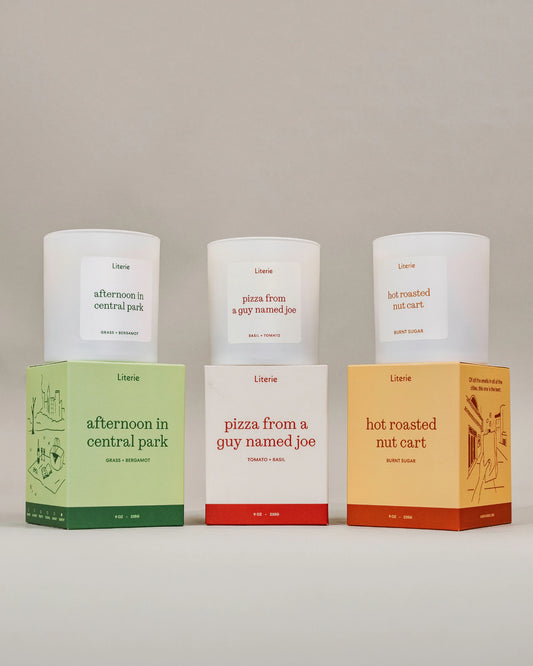 Three Literie scented candles in white glass with white labels, displayed alongside their colorful cardboard boxes on a neutral studio background. The boxes are colored light green, white with red, and light orange, matching the scents: 'afternoon in central park', 'pizza from a guy named joe', and 'hot roasted nut cart'.