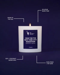 A lit Literie candle in a white jar on a blue  background, surrounded by white callout lines and text highlighting product features: 'Made in the USA', '50 Hour Burn Time', 'Soy + Coconut Blend Wax', 'Non-Toxic + Pet Safe', and the scent 'i ran the tcs new york city marathon'.
