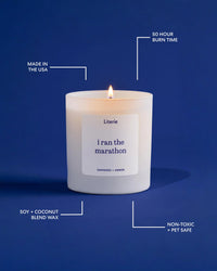 A lit white Literie candle in a matte white glass jar, centered on a dark blue background with white callout lines and text highlighting product features: 'Made in the USA,' '50 Hour Burn Time,' 'Soy + Coconut Blend Wax,' and 'Non-Toxic + Pet Safe.' The name 'i ran the marathon' is visible on the front label.