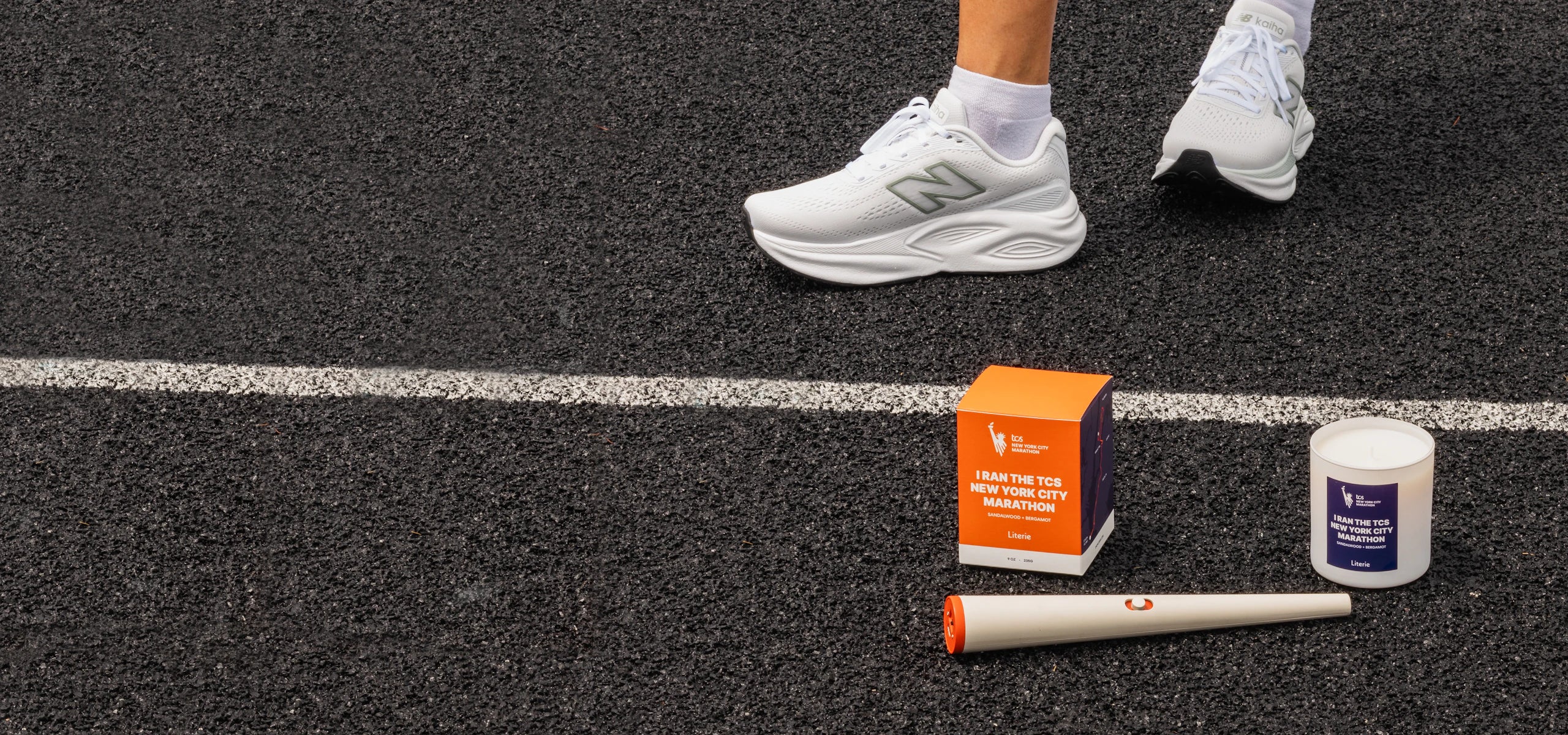 A Literie candle next to its orange and navy blue box on a running track. The candle label reads 'I Ran The TCS New York City Marathon' and the box shows the TCS logo.