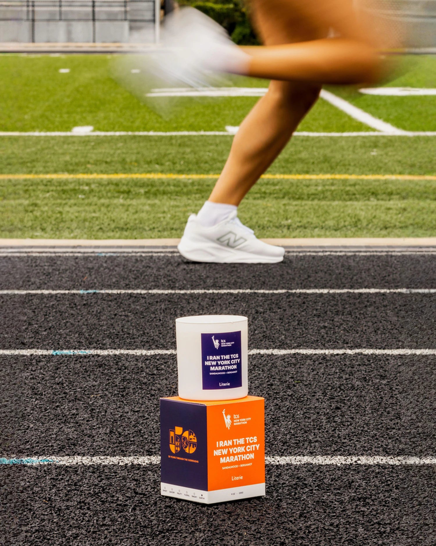 The NYC Marathon candle and box is placed on a running track, with a blurred runner's legs in the background on an athletic track.