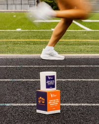 The NYC Marathon candle and box is placed on a running track, with a blurred runner's legs in the background on an athletic track.