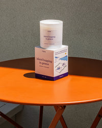 "Island Hopping in Greece" candle and packaging on an orange table with a gray background.