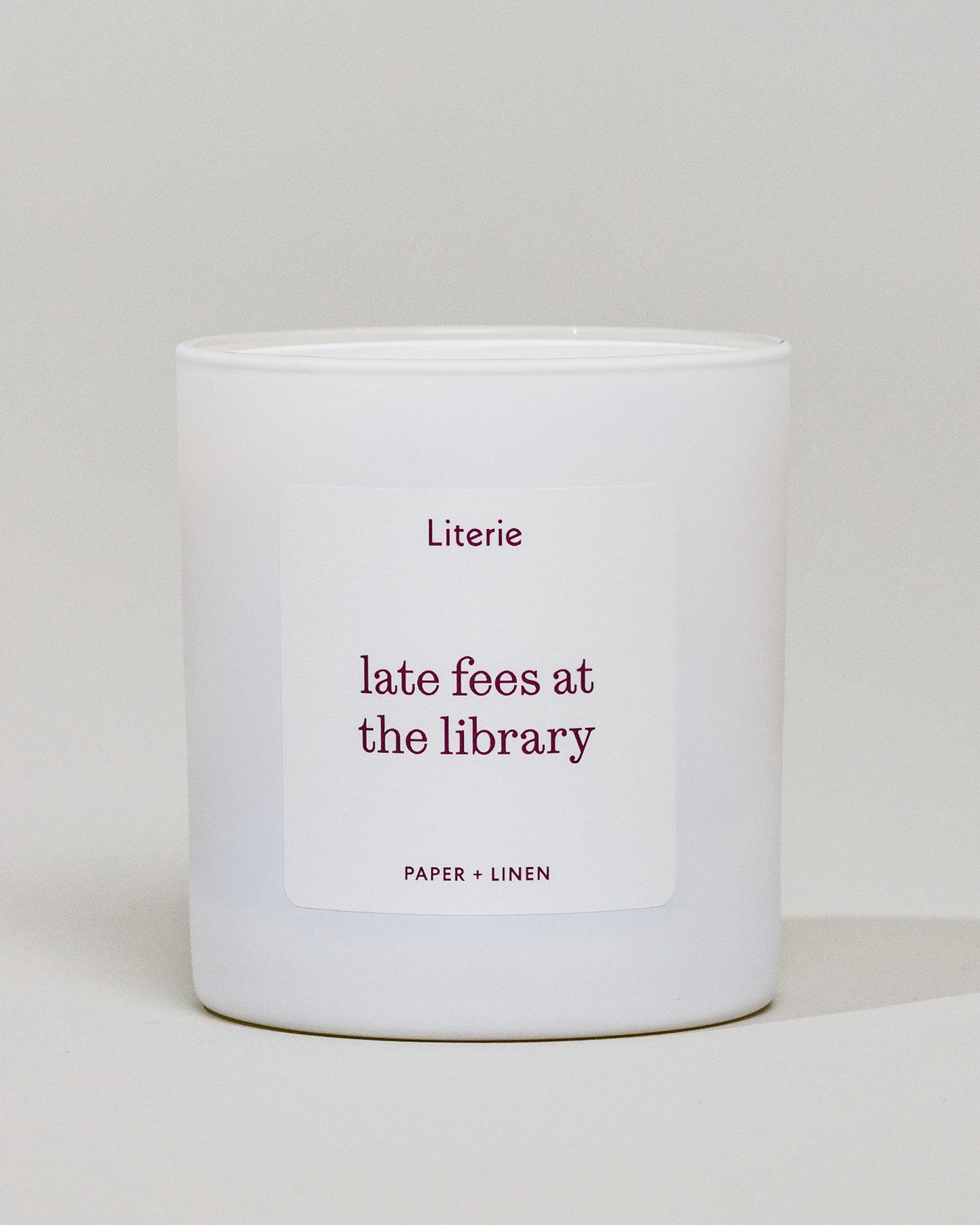 A white scented candle in a cylindrical glass jar with a white label that reads 'Literie late fees at the library' and 'PAPER + LINEN' in burgundy text, set against a neutral light gray background.
