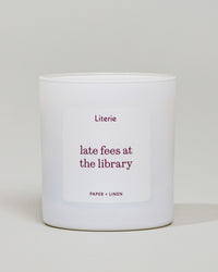 A white scented candle in a cylindrical glass jar with a white label that reads 'Literie late fees at the library' and 'PAPER + LINEN' in burgundy text, set against a neutral light gray background.