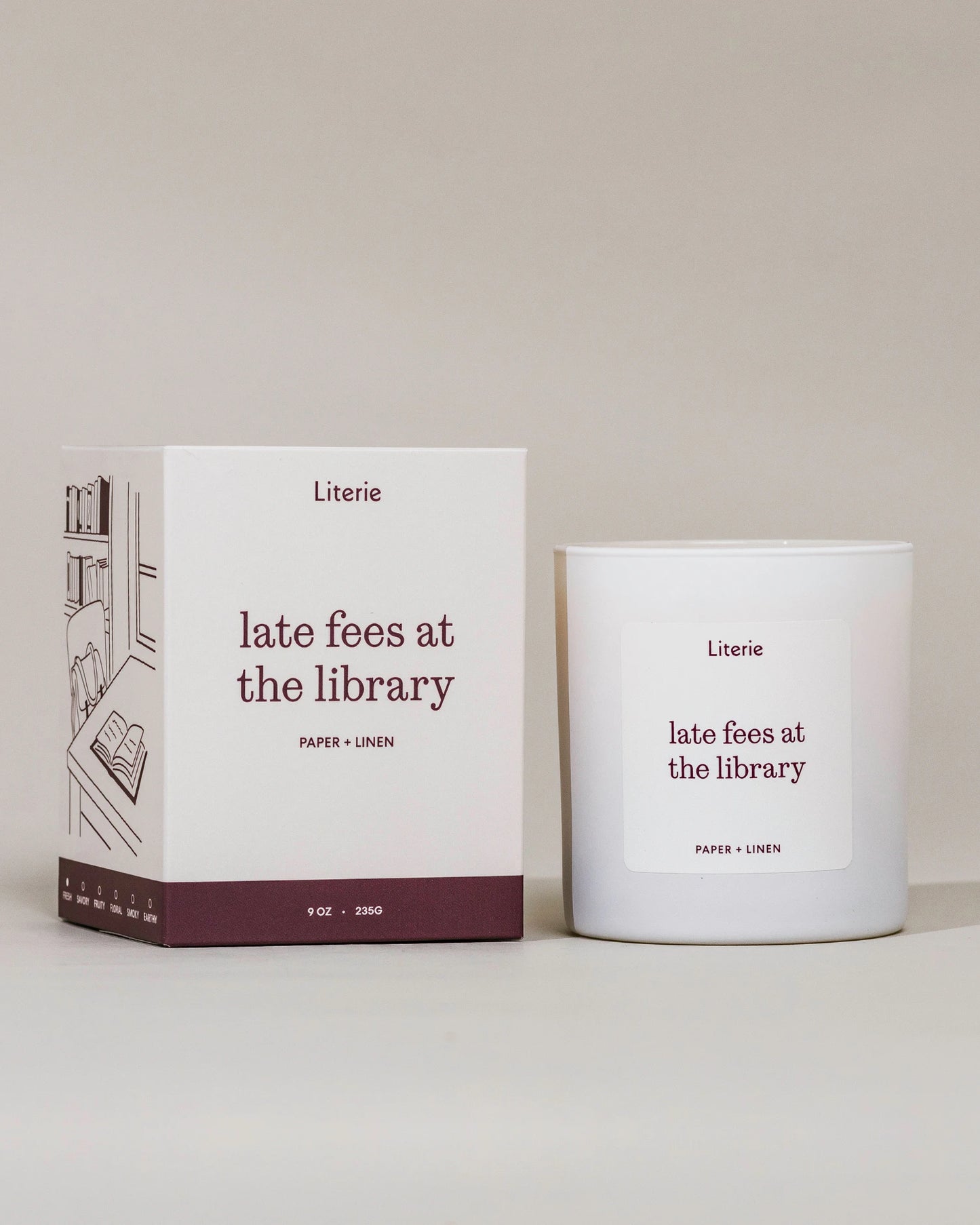 A white candle jar and its matching white box with dark burgundy text and illustration, labeled Literie 'late fees at the library' on a warm off-white background.