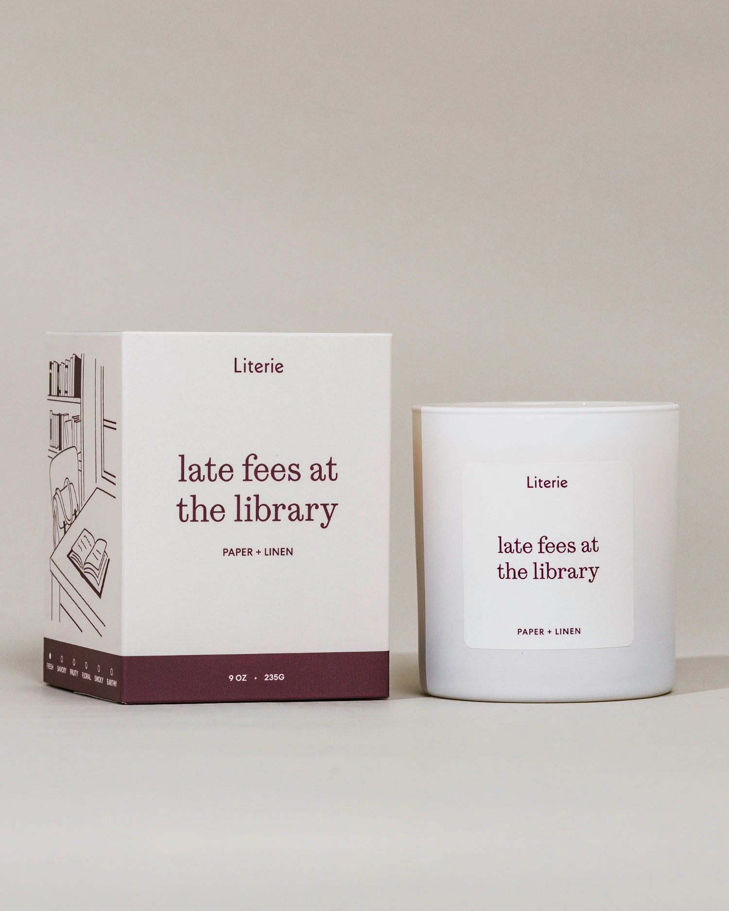 A white candle jar and its matching white box with dark burgundy text and illustration, labeled Literie 'late fees at the library' on a warm off-white background.