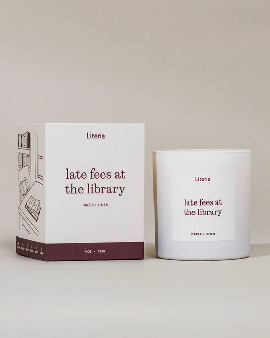 A white candle jar and its matching white box with dark burgundy text and illustration, labeled Literie 'late fees at the library' on a warm off-white background.