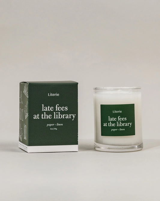 A 2.5oz candle in a clear glass jar with a dark green label, standing next to its matching dark green packaging box on a light gray background. The label and box are printed with the brand name 'Literie' and the scent name 'late fees at the library'.