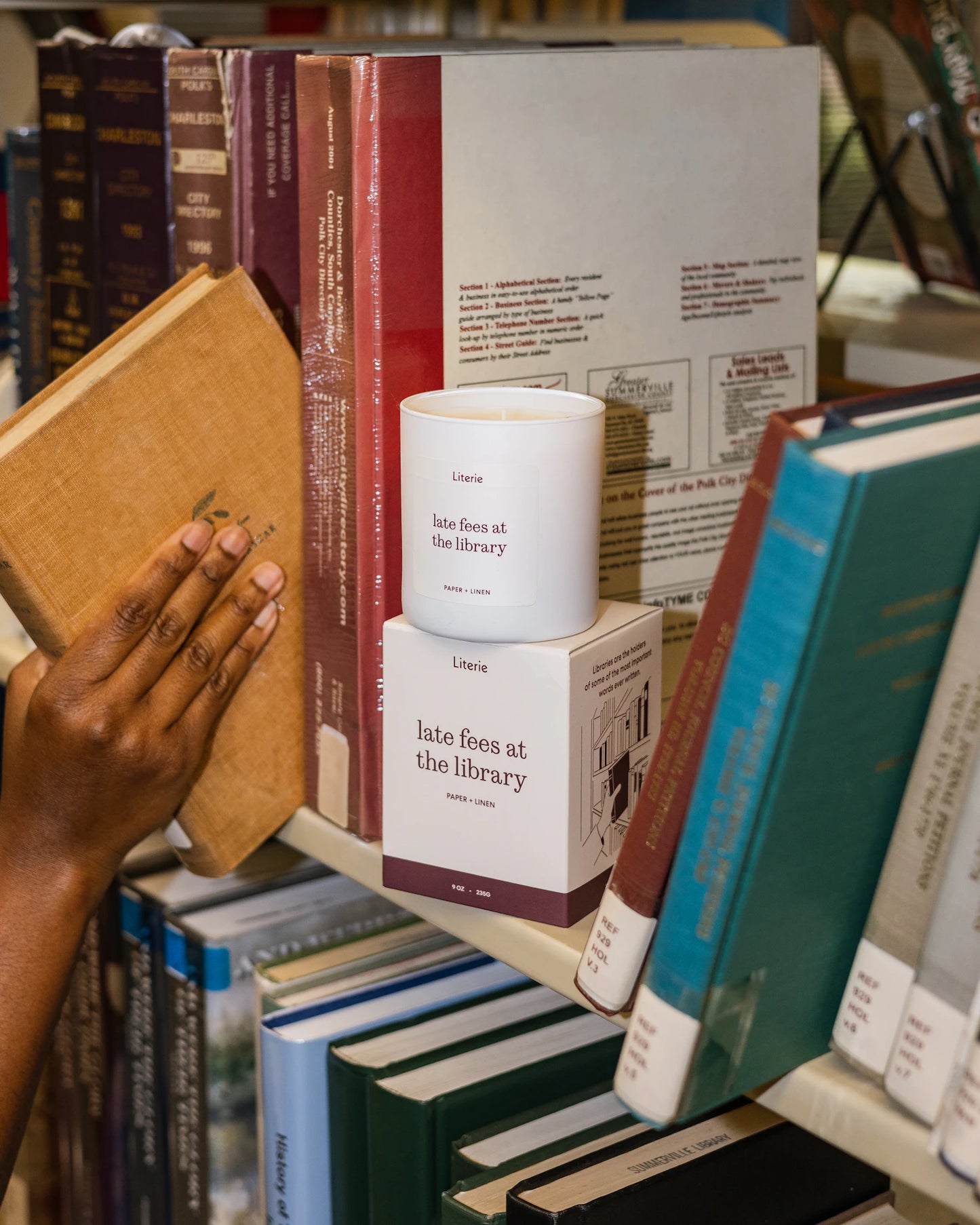 A white candle from the brand Literie sits on top of its box on a bookshelf, styled next to a person's hand pulling out a book.