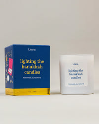 A white glass candle jar and its blue and gold illustrated packaging box, both labeled 'Literie lighting the hanukkah candles' and 'Powdered Jelly Donuts,' on a clean light gray studio background.