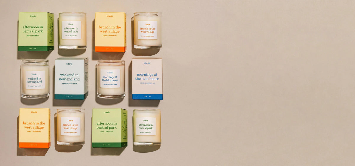A grid candles in clear glass jars with colorful paper labels and matching colored packaging boxes, arranged on a light beige background. The labels feature text such as 'afternoon in central park' and 'weekend in new england'.