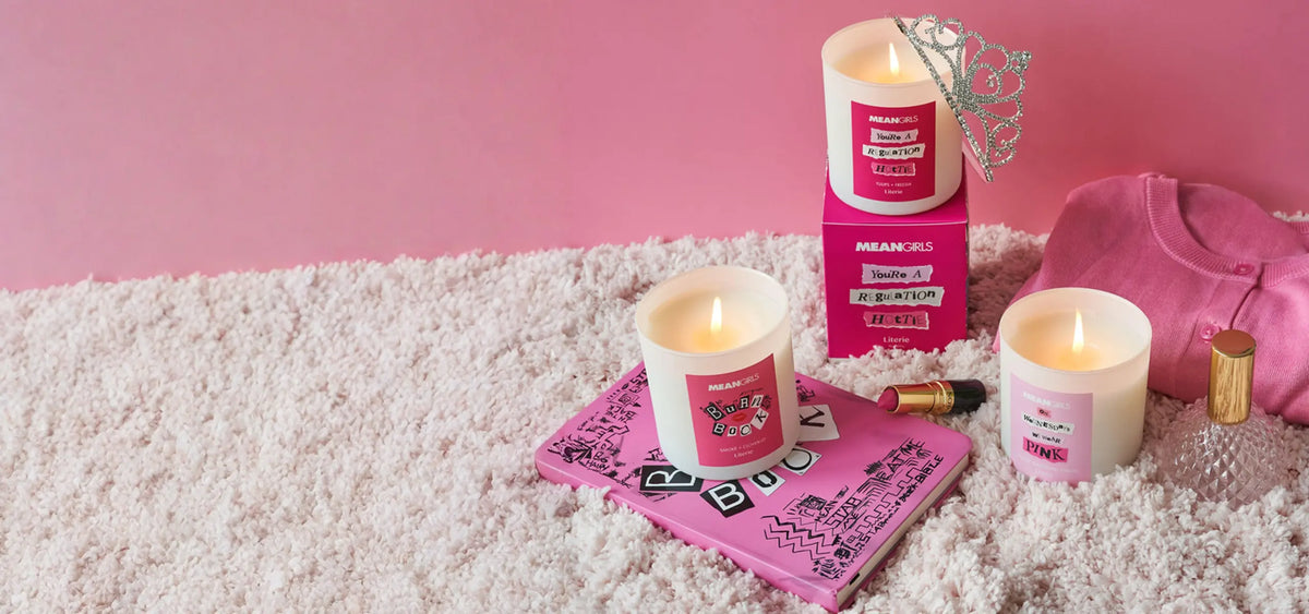 Three Mean Girls scented candles by Literie in white glass jars with pink labels, arranged on a white shag rug with pink props including a tiara, notebook, lipstick, perfume bottle, and pink sweatshirt against a pink background.