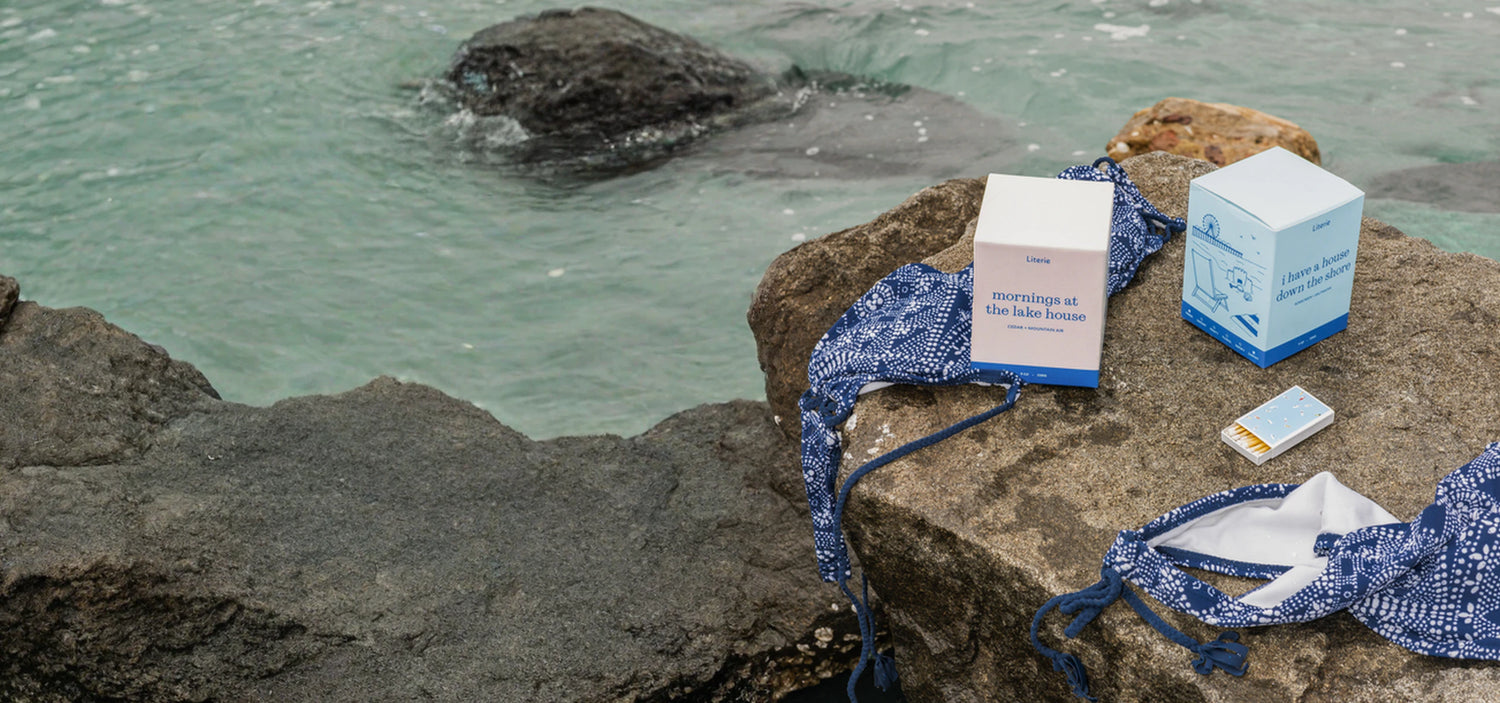 Blue swimwear and Literie candle boxes on rocks by a body of water.