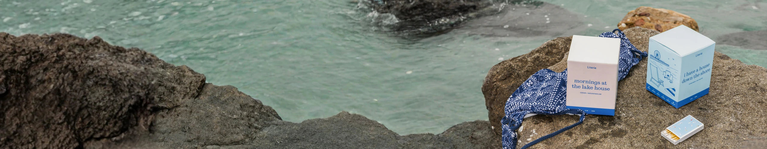 Two Literie candle boxes on a rocky surface near water