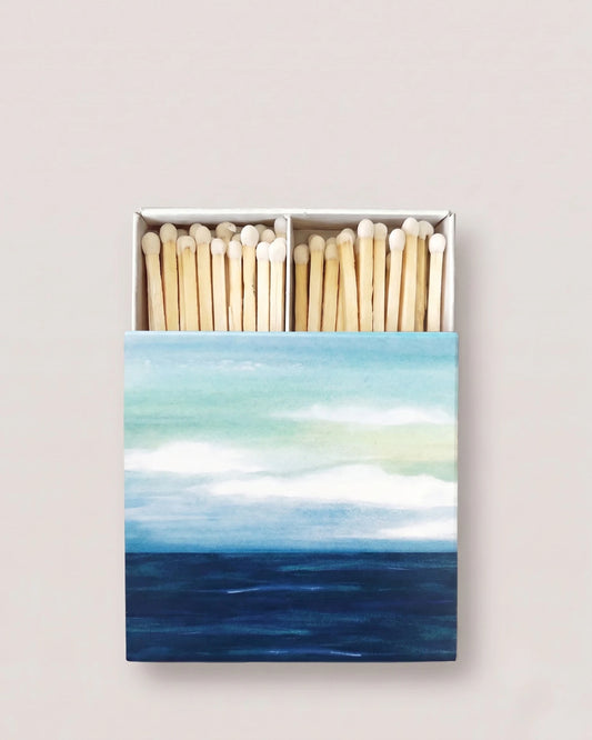 An open matchbox with natural wood matches inside, featuring a painted ocean scene on the front cover in shades of blue, teal, and white, set against a solid light beige background.