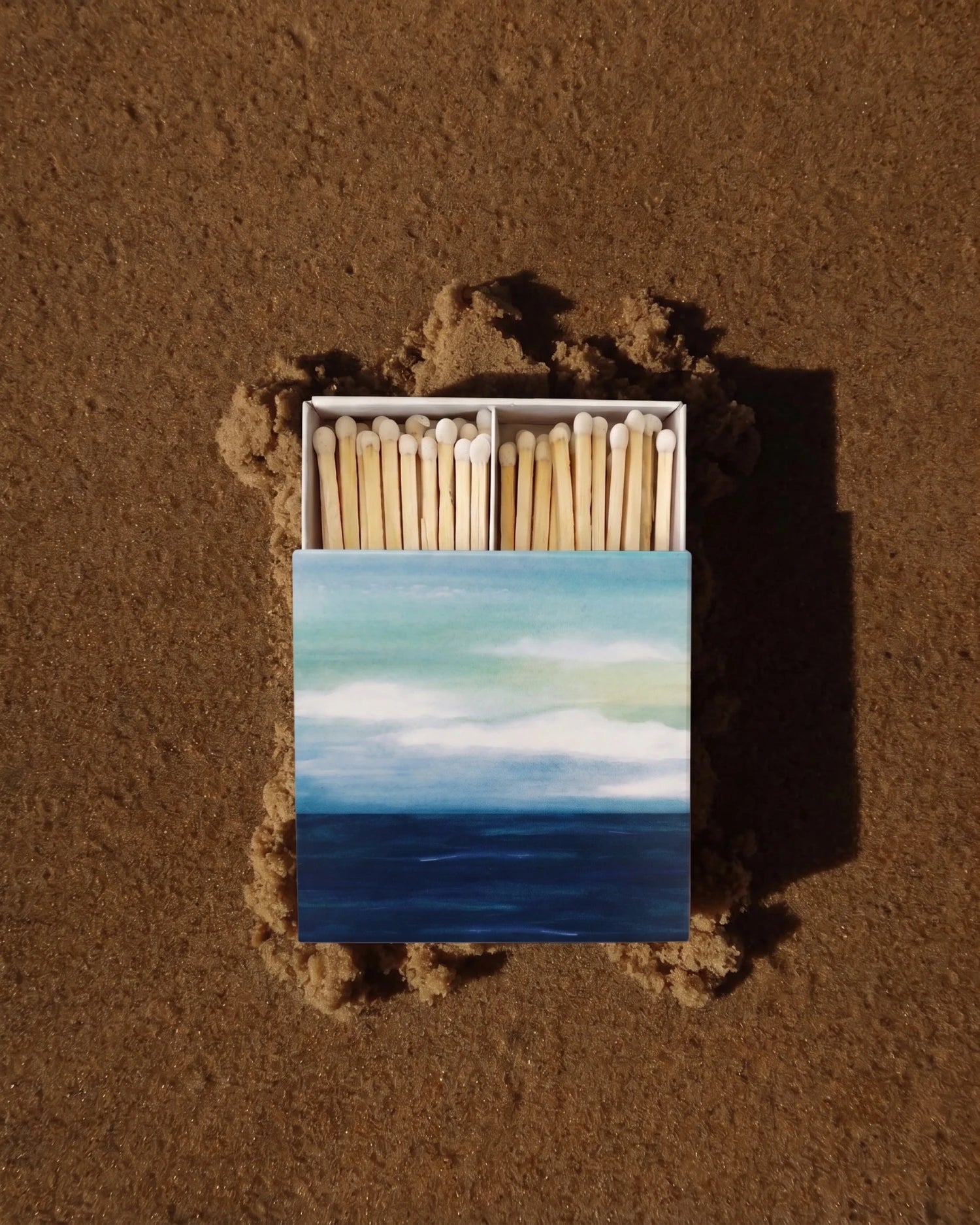 An open matchbox with painted ocean and sky artwork on the cover, placed on sand with a few matches visible. The product is shown in a real outdoor setting.