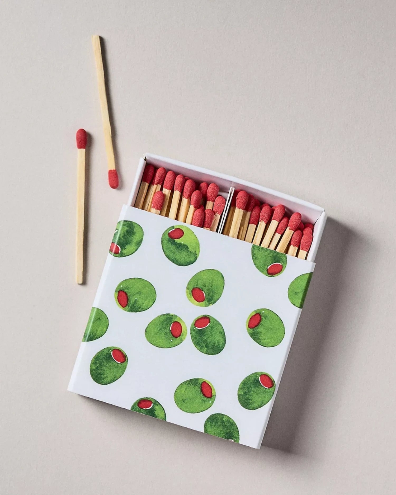 A box of matches with a white lid printed with green olives and red peppercorns, shown open with wooden matches and red tips visible, on a light beige background.