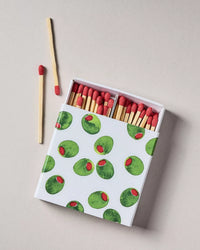 A box of matches with a white lid printed with green olives and red peppercorns, shown open with wooden matches and red tips visible, on a light beige background.