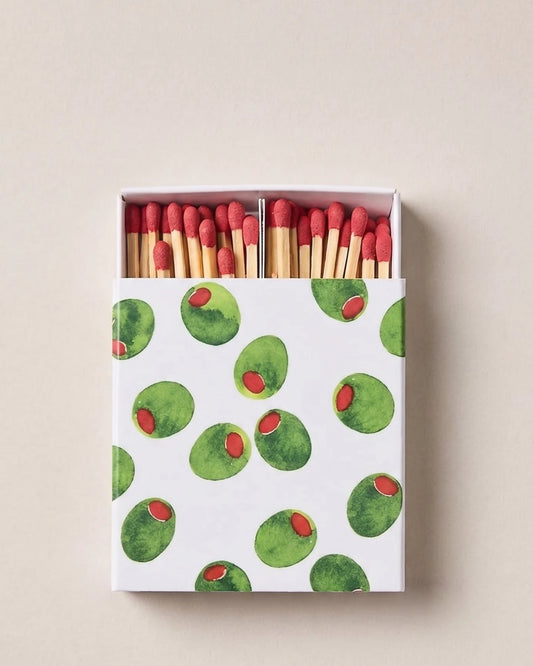 A box of matches with a white cover decorated with a pattern of green olives and red peppercorns, shown open to reveal red-tipped wooden matches against a neutral beige background.