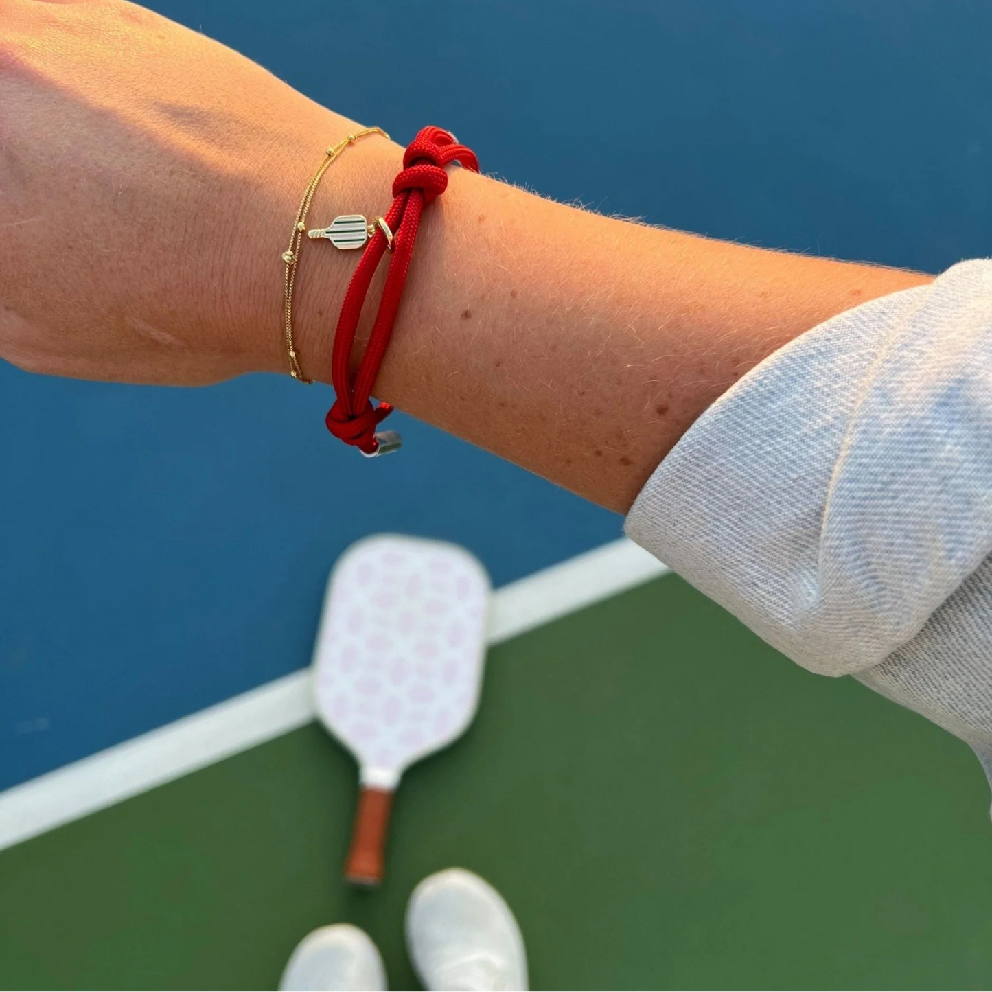 Person's arm with a red bracelet and gold bracelet with charm on a pickleball court.