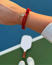 Person's arm with a red bracelet and gold bracelet with charm on a pickleball court.