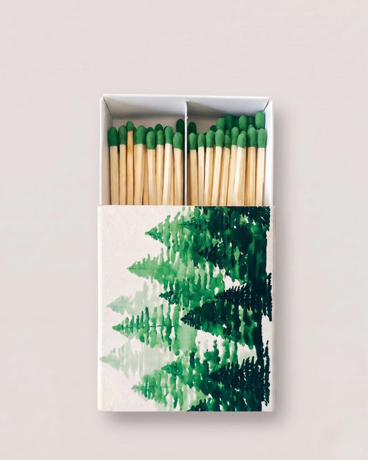 A matchbox with a watercolor-style green pine forest design on the cover, shown open with natural wood matches and green-tipped matches inside, against a light beige background.