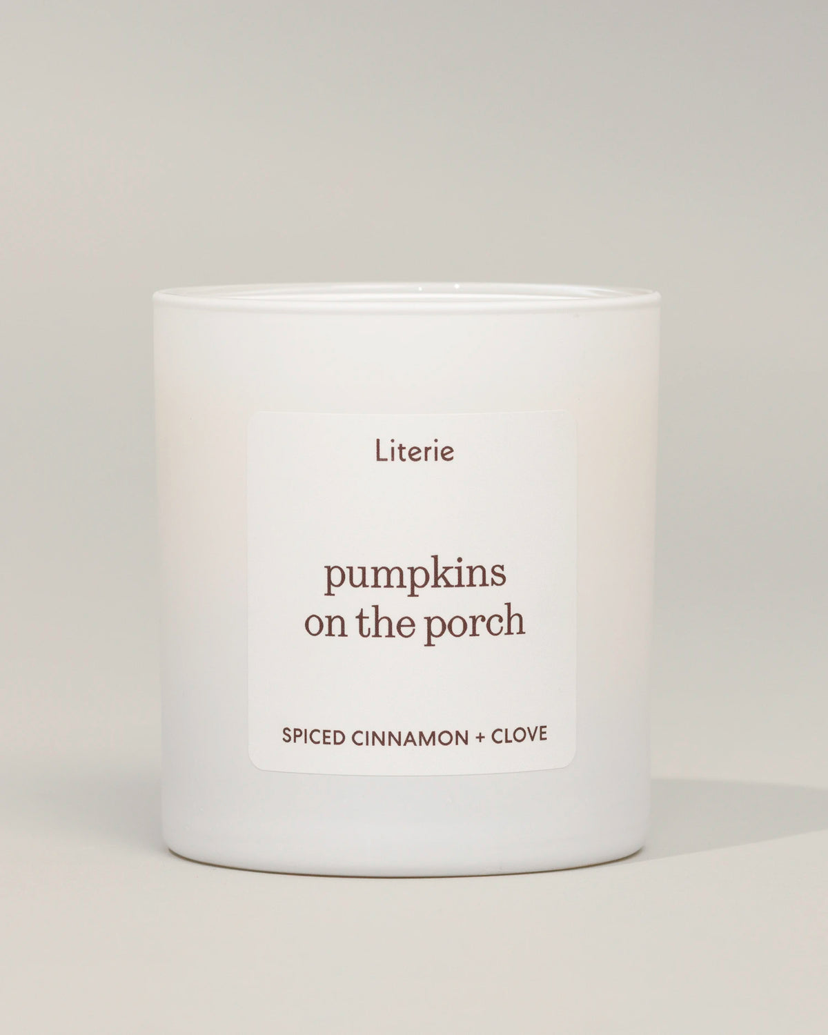 A white glass jar candle with a minimalist white label reading 'Literie pumpkins on the porch' and 'SPICED CINNAMON + CLOVE' in brown text, photographed against a neutral light gray background.