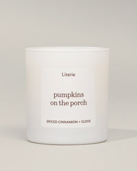 A white glass jar candle with a minimalist white label reading 'Literie pumpkins on the porch' and 'SPICED CINNAMON + CLOVE' in brown text, photographed against a neutral light gray background.