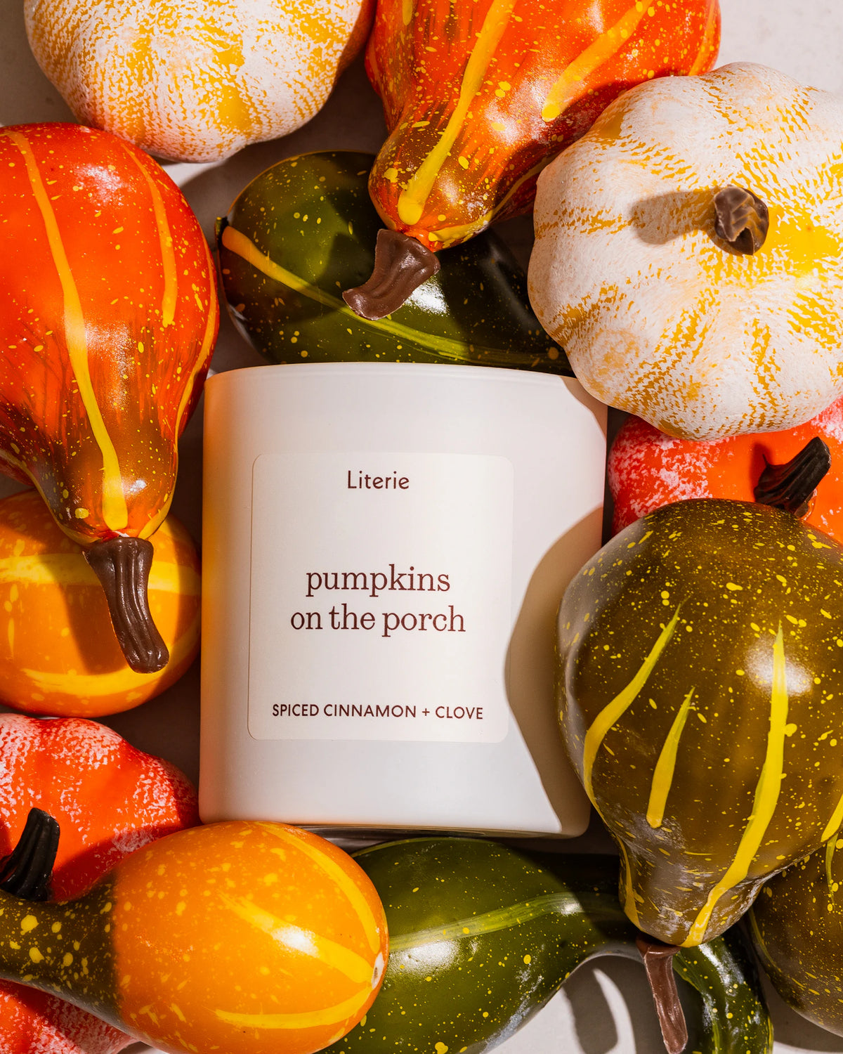 A white candle labeled 'Literie pumpkins on the porch' surrounded by decorative orange, yellow, and green gourds in a styled flat-lay arrangement.