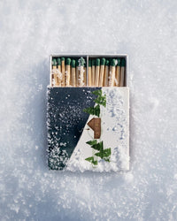A decorative matchbox filled with wooden matches and green-tipped matches, placed on a snowy surface. The matchbox lid features a winter scene with a small house, evergreen trees, and a dark blue sky.