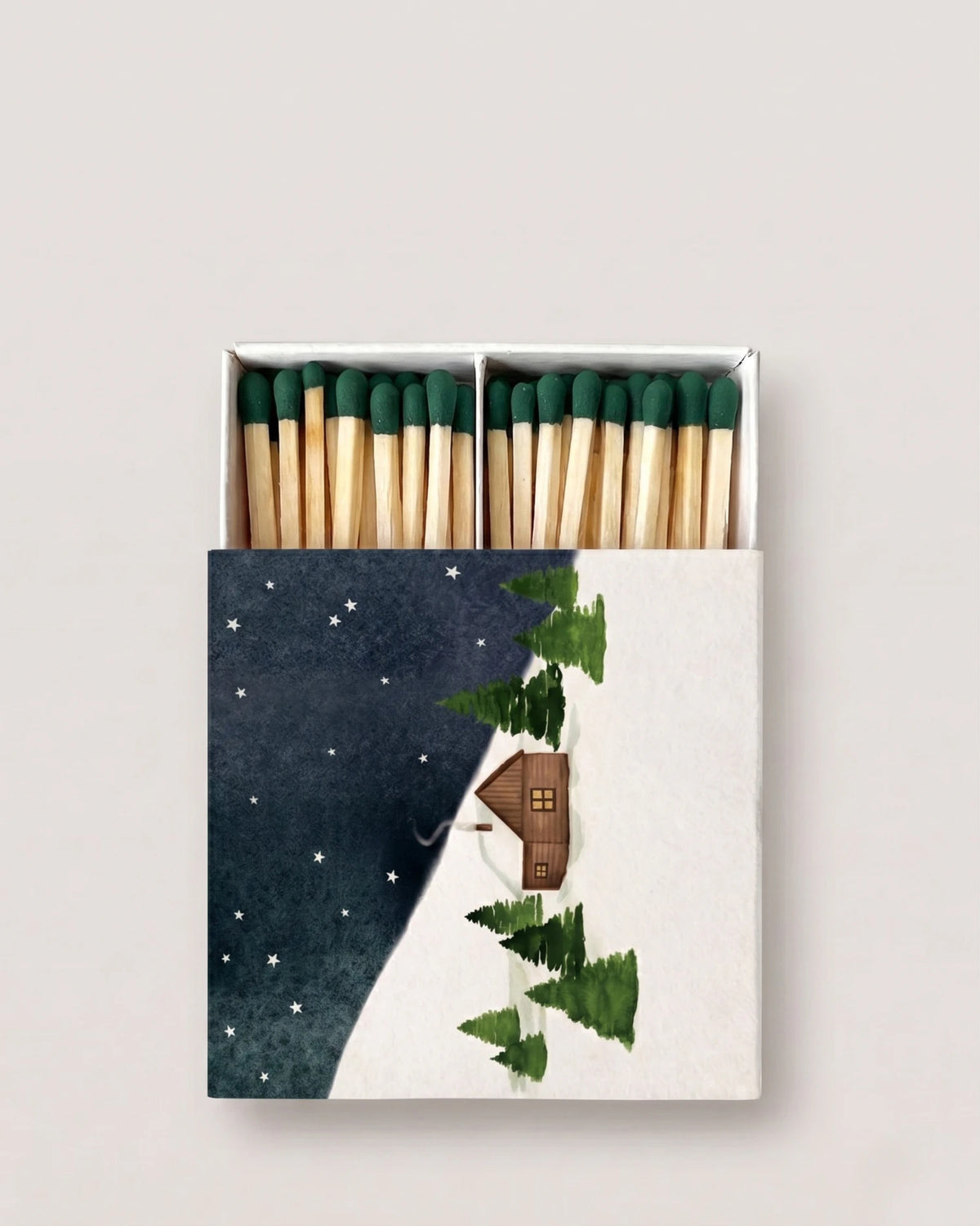 A matchbox of wooden matches with dark green tips, featuring a winter night illustration on the front with a small cabin, evergreen trees, and a starry dark blue sky.