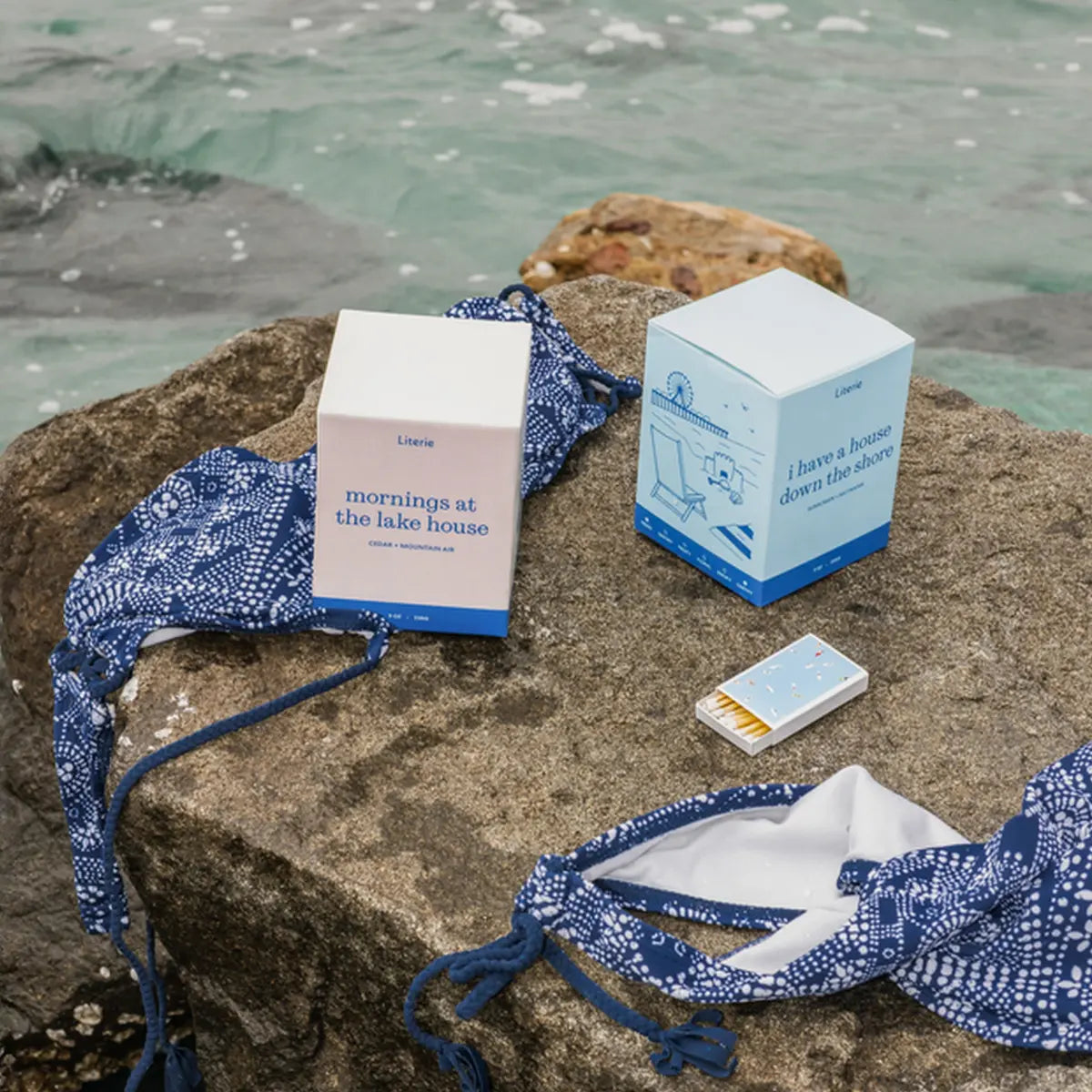 Two Literie candle boxes and a small matchbox styled on a large rock with blue-and-white patterned swimwear and water in the background.