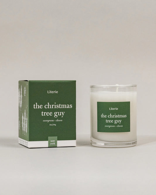 A 2.5oz candle in a clear glass jar next to its dark green packaging box, both featuring the brand name 'Literie' and the scent name 'the christmas tree guy' on a neutral light gray background.