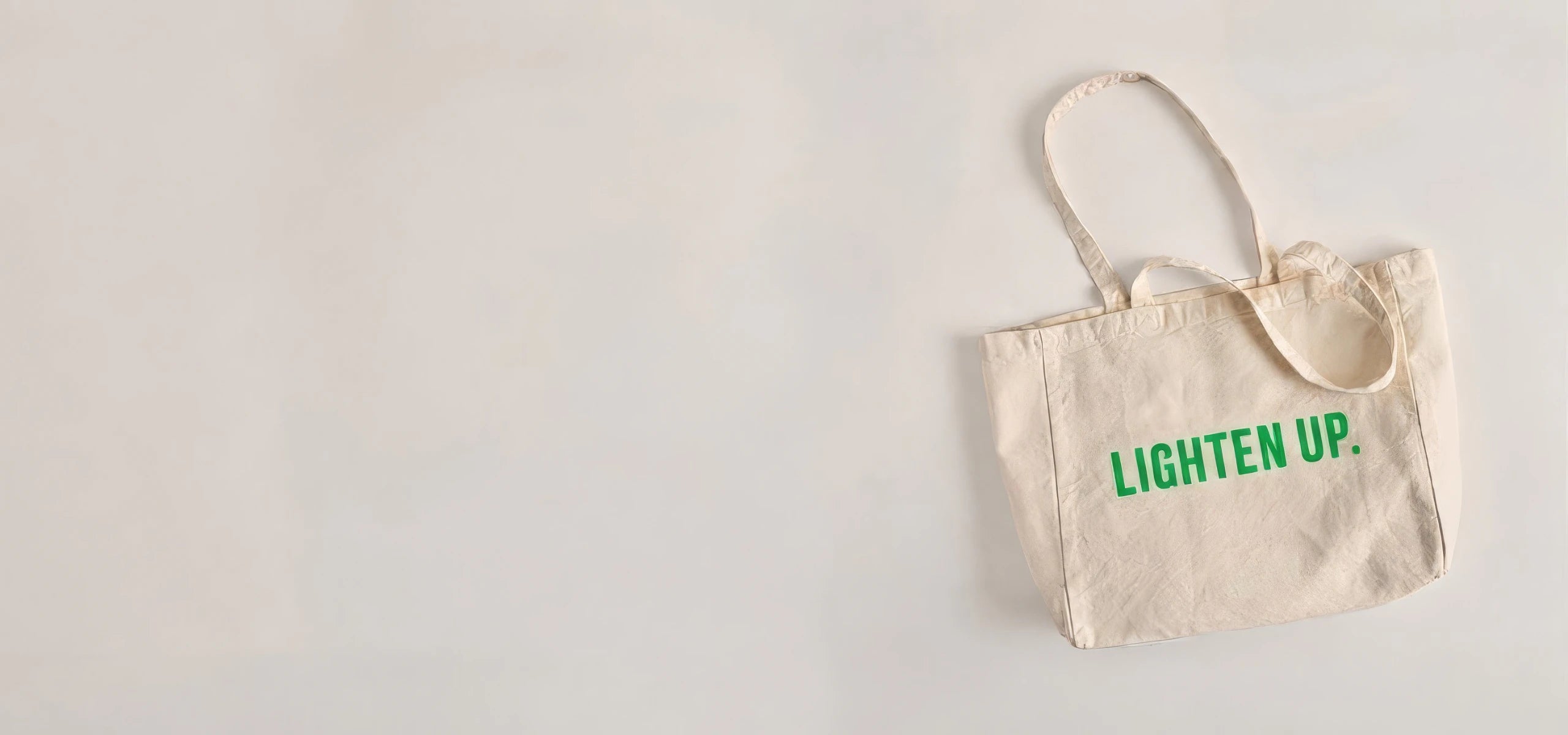 A natural canvas tote bag with green text reading 'LIGHTEN UP.' on the front, photographed against a plain off-white background with ample negative space on the left.
