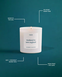 A lit Literie candle in a white jar on a solid orange background, surrounded by white callout lines and text highlighting product features: 'Made in the USA', '50 Hour Burn Time', 'Soy + Coconut Blend Wax', 'Non-Toxic + Pet Safe', and the scent 'weekend in new england'.