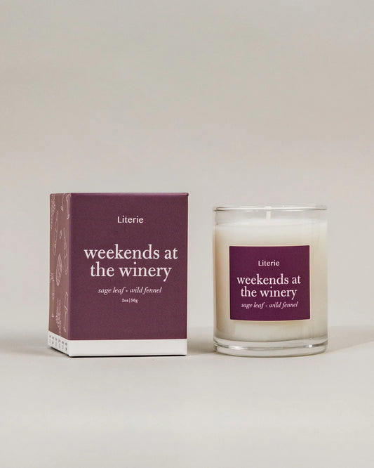 A Literie 'weekends at the winery' scented little candle in a clear glass jar next to its matching dark purple packaging box, set against a neutral light gray background.