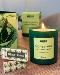 A lit WICKED 'defying gravity on broadway' candle in a dark green glass jar sits on a wooden table next to its patterned packaging box and two wrapped matches.