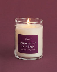 A lit 2.5oz candle in a clear glass jar with a purple label reading 'Literie weekends at the winery' on a matching purple background.